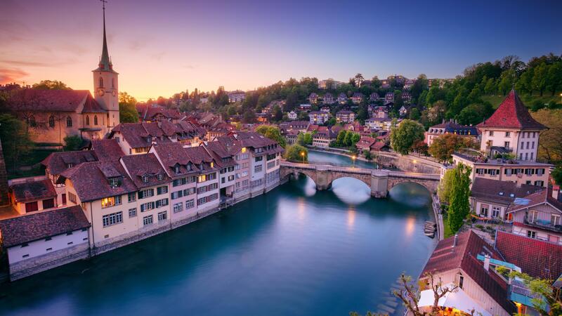 Charming town by a river at dusk, with a bridge, rooftops, and a church spire under a colorful sky.