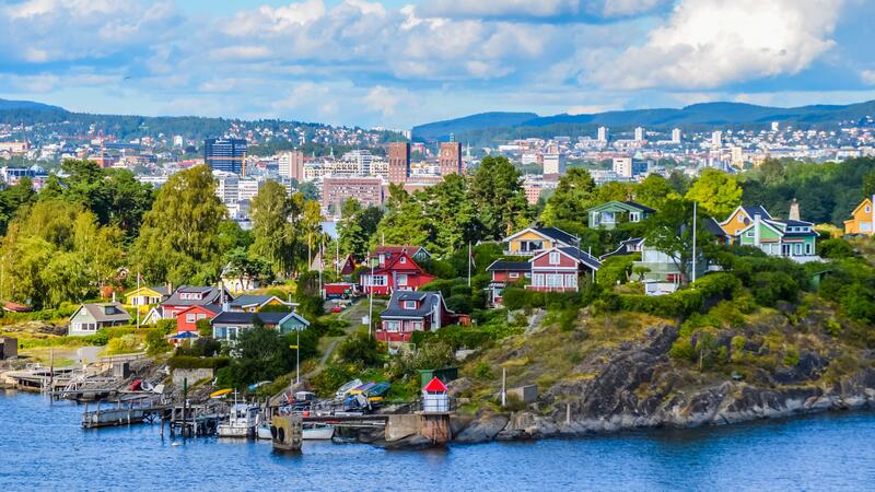 Colorful houses on a lush hillside by the sea, cityscape visible in the background, under a partly cloudy sky.